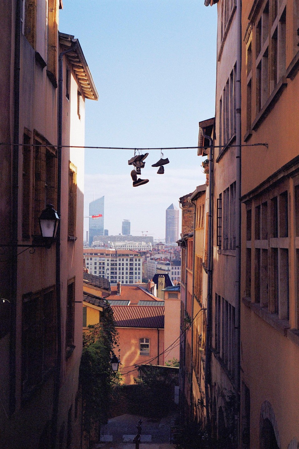 A pair of shoes hangs from a wire above a narrow street with buildings on either side and a city skyline in the distance.
