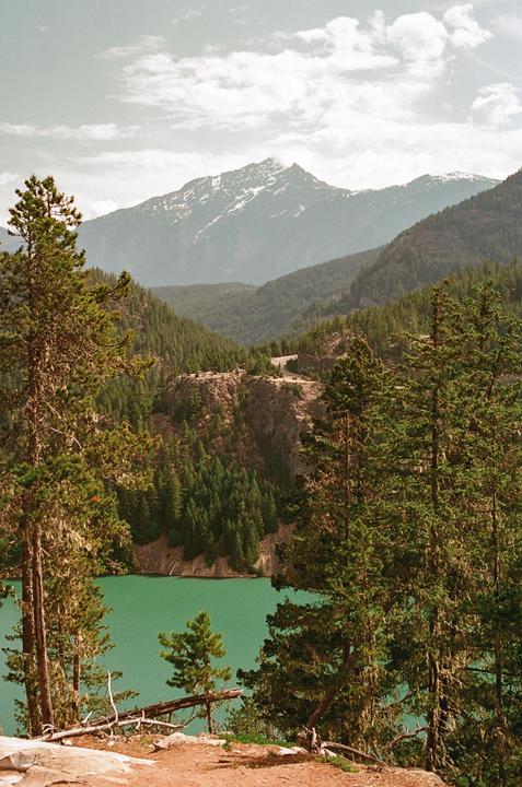 A vibrant green lake is nestled among a dense forest with snow-capped mountains in the distance under a cloudy sky.