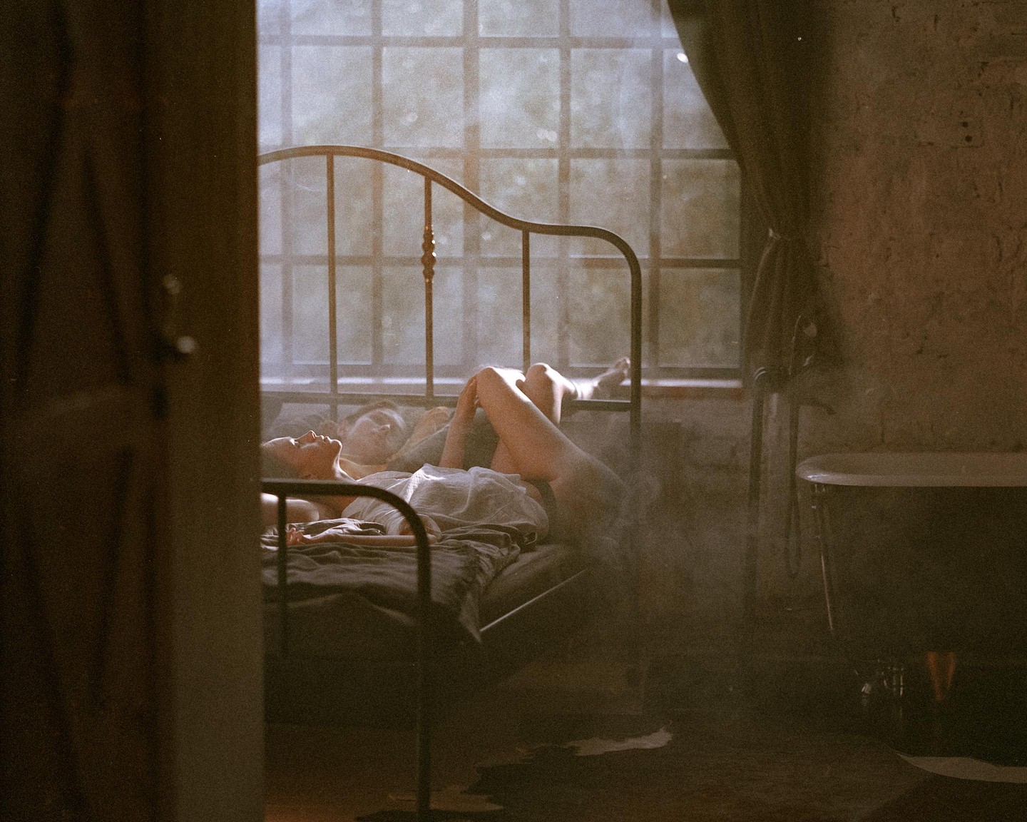A woman lies on a bed in a hazy room with a window and a bathtub.