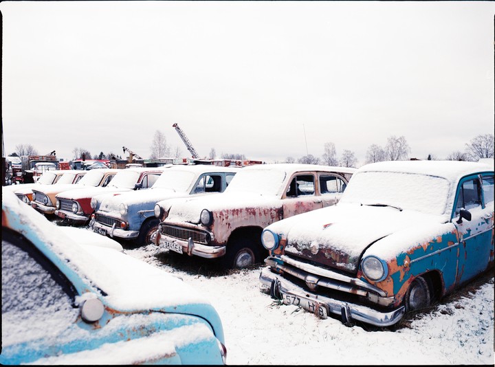 A line of snow-covered, rusty vintage cars sits in a field under a white sky.