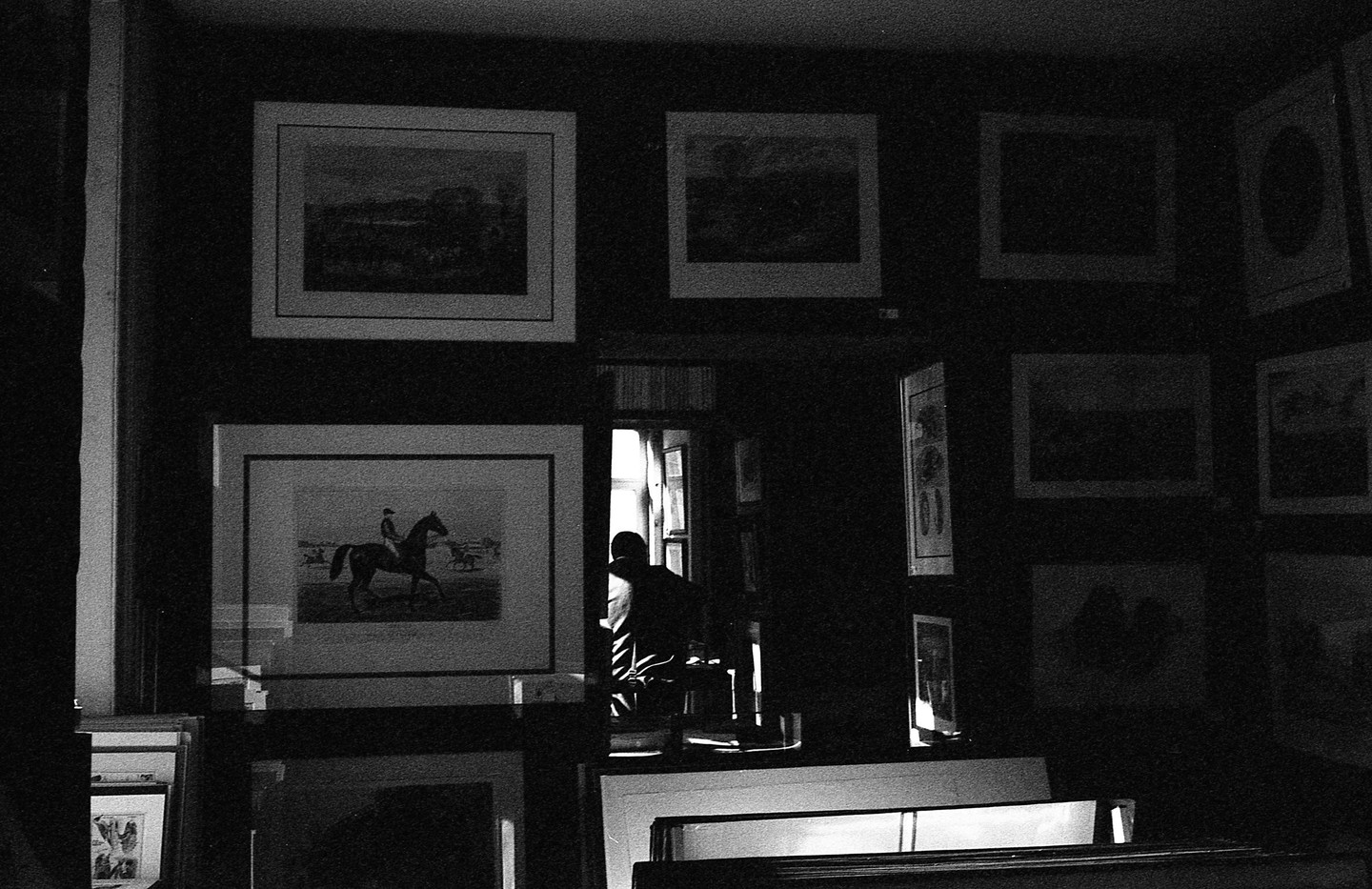 A dark room filled with framed pictures on the walls and stacked below, with a person silhouetted in a doorway in the background.