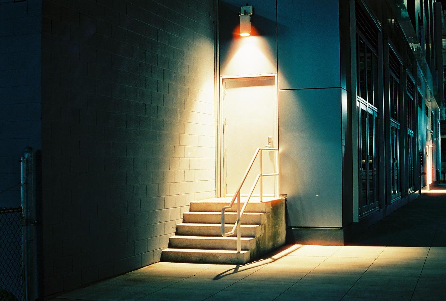 A concrete staircase with a white railing leads to a door under a bright light at night.