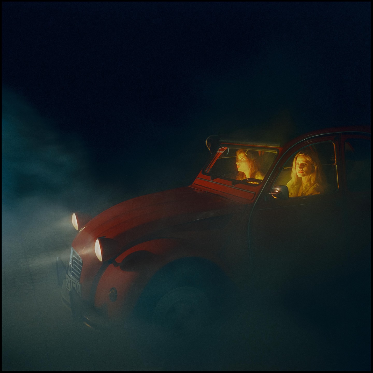 Two women are in a red car at night, surrounded by fog.