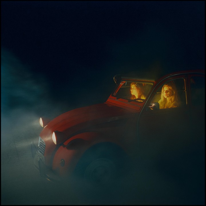 Two women are in a red car at night, surrounded by fog.