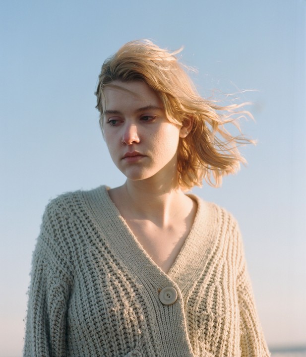 A woman with blonde hair and a knitted cardigan looks down against a clear sky.