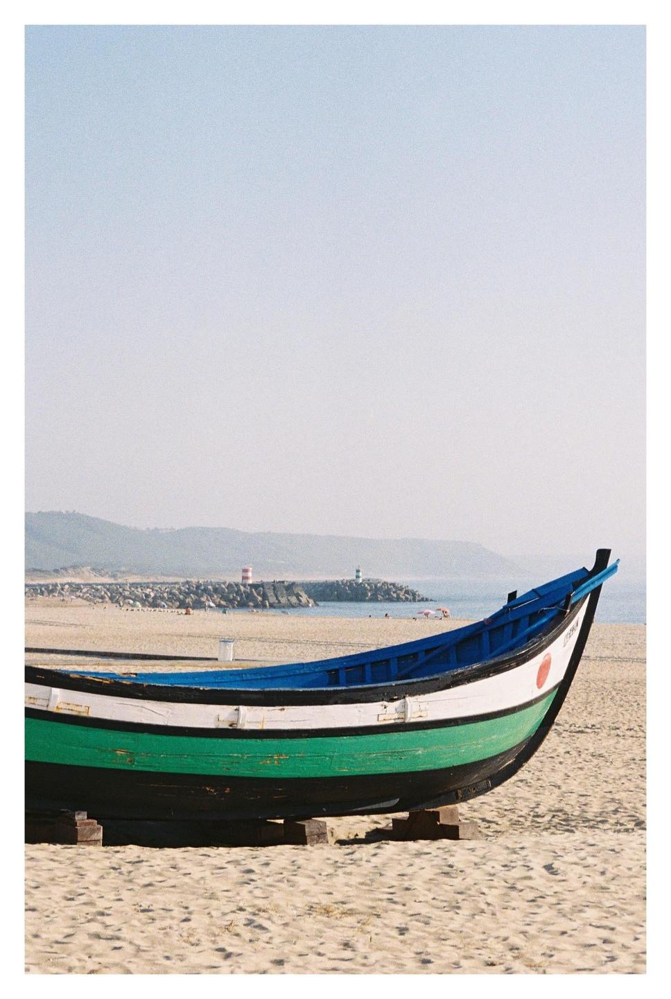 A colorful boat rests on a sandy beach with a distant coastline and lighthouses under a clear sky.