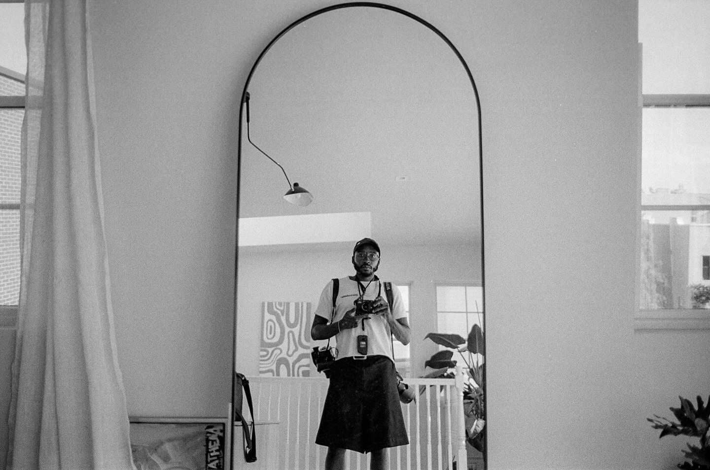 A man wearing a hat and glasses takes a self-portrait in a large arched mirror.