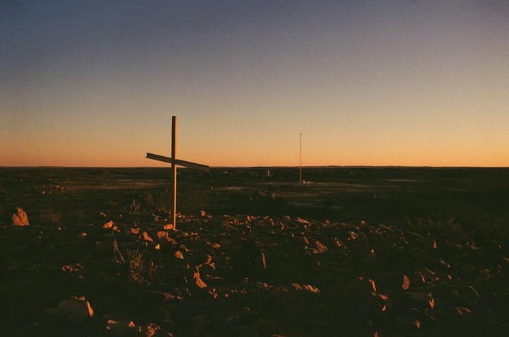 A cross stands on a rocky, barren landscape under a gradient sky at sunset.