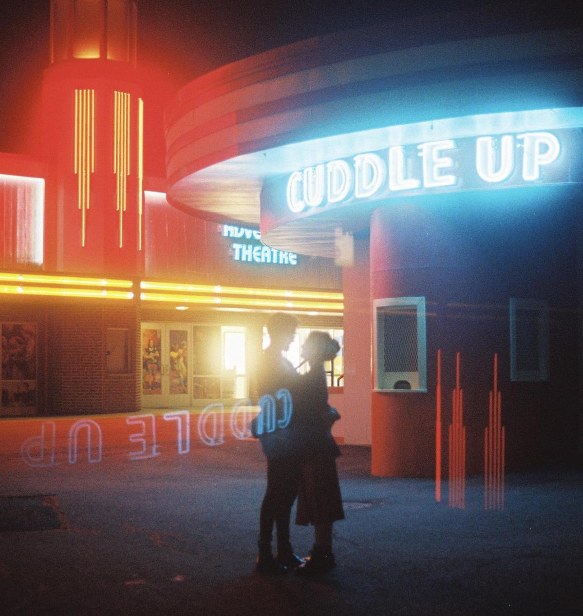A couple embraces in front of a movie theater with neon signs that read "Cuddle Up" and "Theatre."
