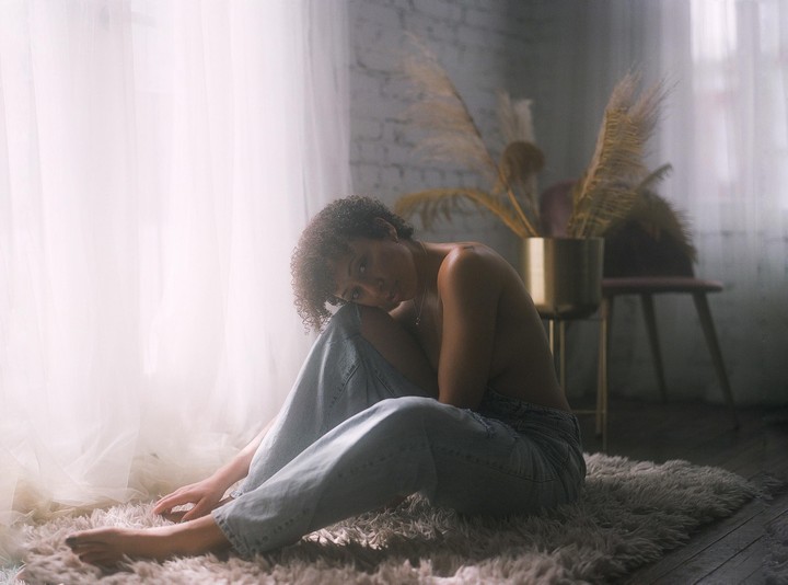 A woman with curly hair sits on a rug in front of a window, looking at the viewer.