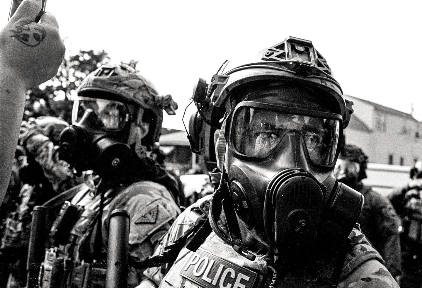 A police officer wearing a gas mask and helmet looks forward while another officer in similar gear stands behind him.