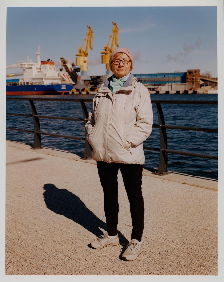 A woman stands on a pier with a body of water and industrial cranes in the background.