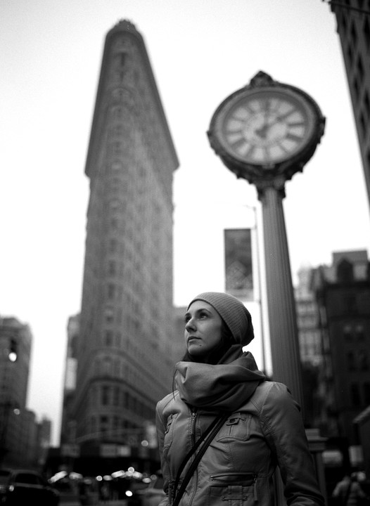 A woman in a beanie and scarf looks up at the Flatiron Building and a large street clock.