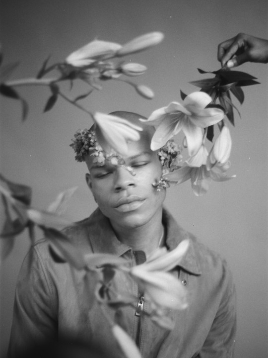 A man with closed eyes has flowers on his head and face, with more flowers in the foreground and a hand holding a branch of leaves in the background.