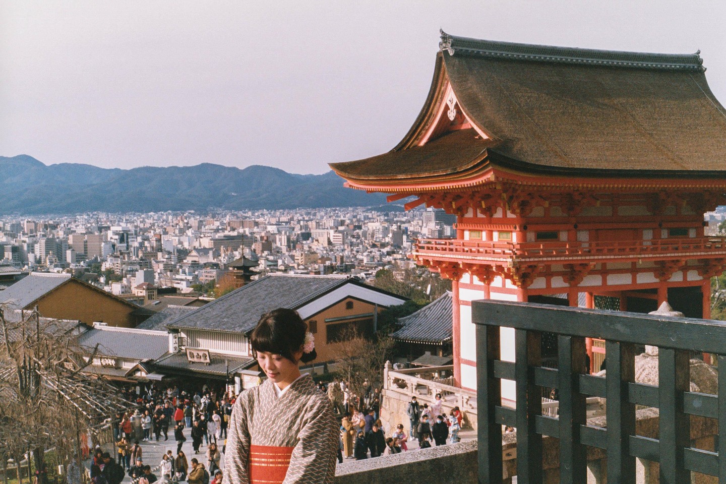 A woman in a kimono stands on a hill overlooking a city with a temple and mountains in the background.