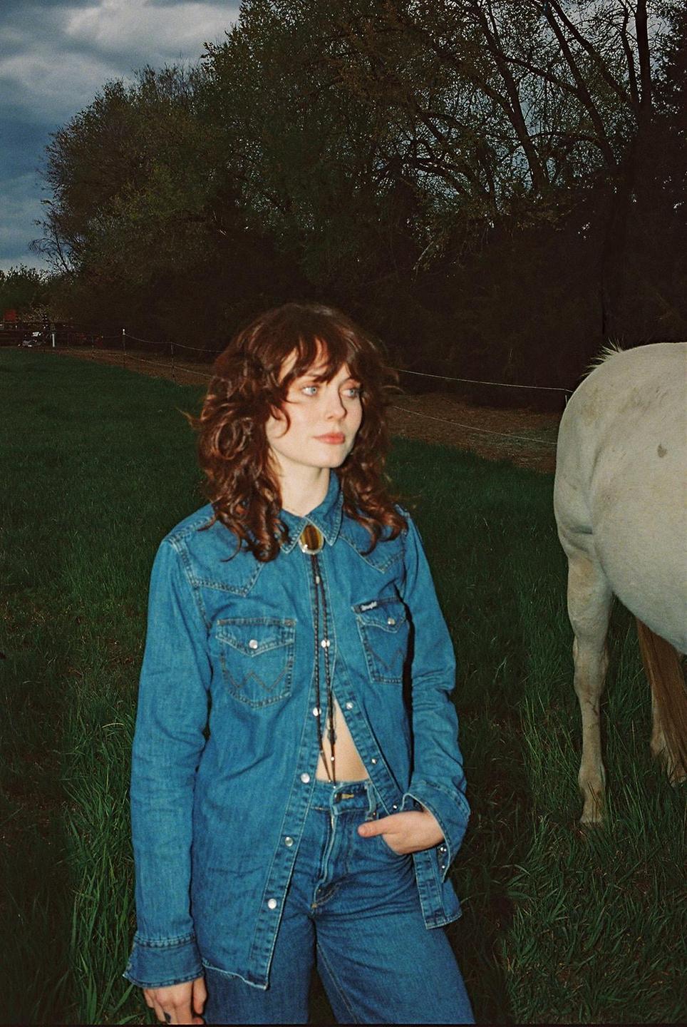 A woman in a denim shirt and jeans stands in a grassy field next to a horse.