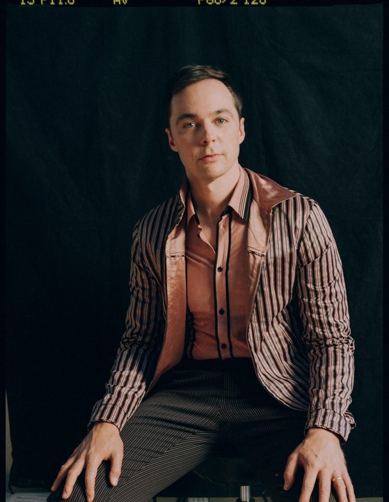 A man in a striped jacket and pink shirt sits against a dark background.