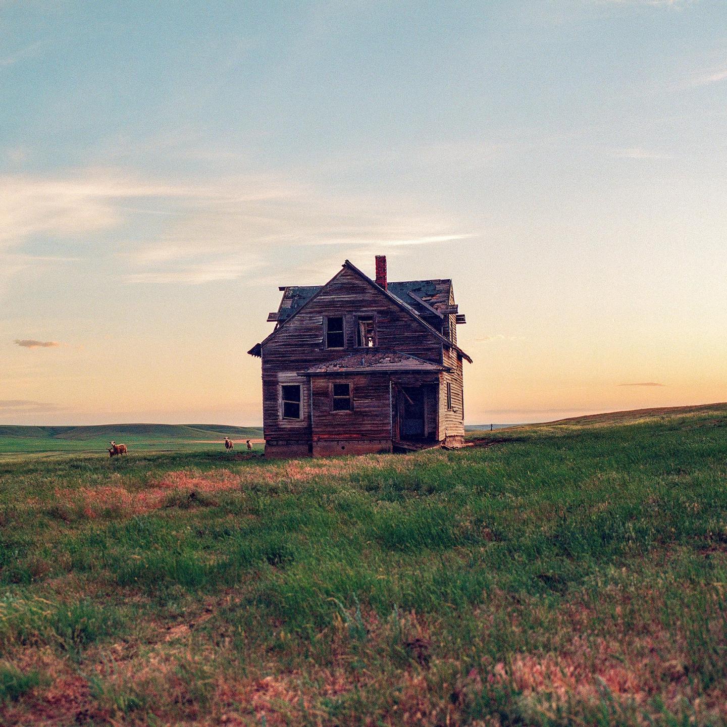 An abandoned wooden house stands in a grassy field under a colorful sky at dusk.