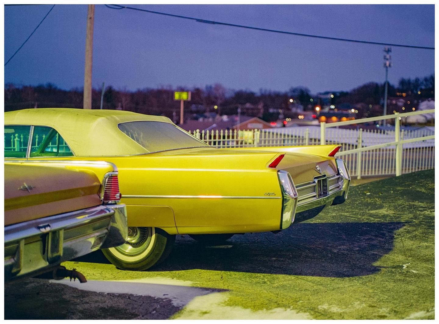 Two vintage cars are parked side by side at night, with a white fence and distant lights in the background.