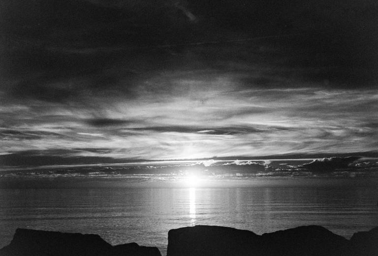 The sun sets over a body of water, casting a bright reflection on the surface, with dark clouds above and silhouetted rocks in the foreground.