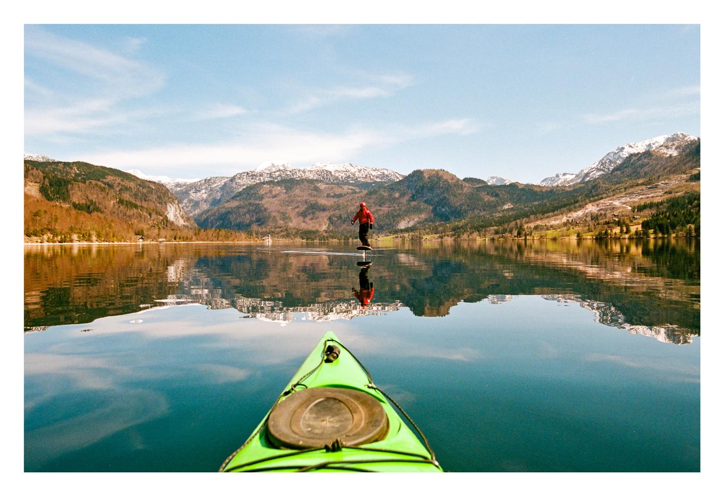 A person on an eFoil is visible from the perspective of a kayak on a lake surrounded by mountains.