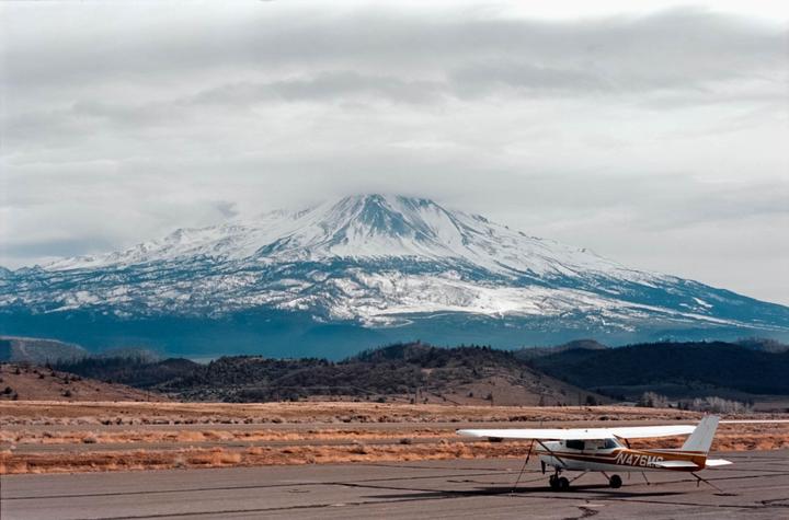 A small airplane is parked on an airfield with a large snow-capped mountain in the background under a cloudy sky.