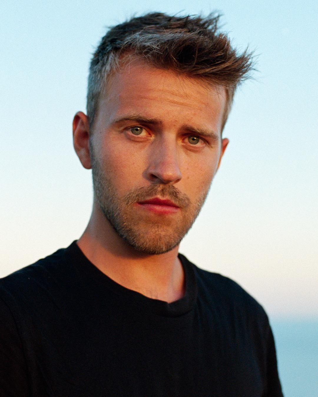 A man with a beard and short hair looks directly at the viewer against a light blue sky.