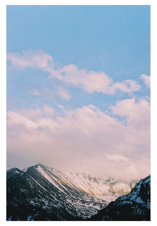 A snow-covered mountain range is partially obscured by clouds under a blue sky.