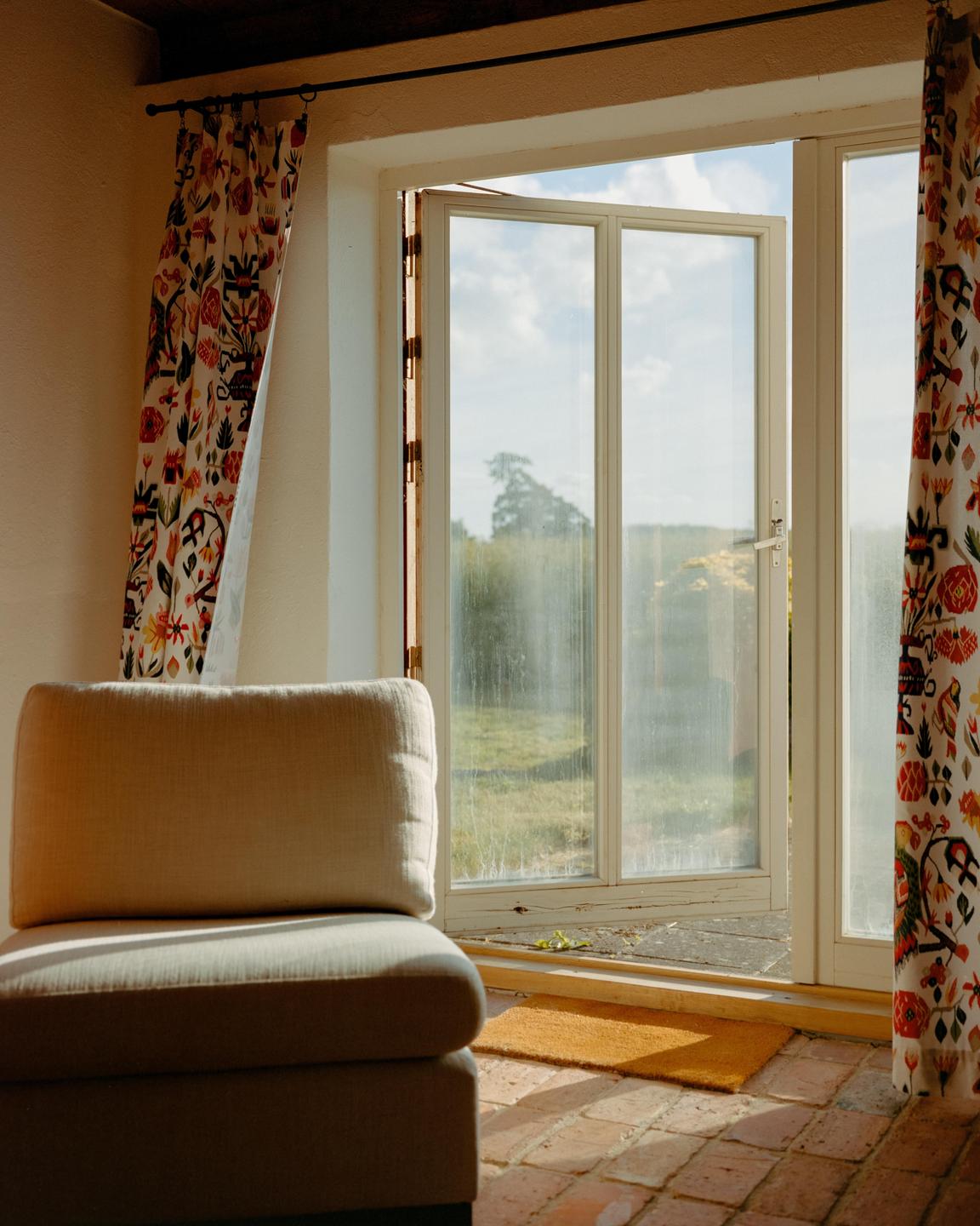 A beige armchair sits on a brick floor next to an open glass door with patterned curtains.