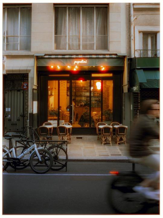 A cafe with outdoor seating and bicycles parked on the street is illuminated by warm light at dusk.