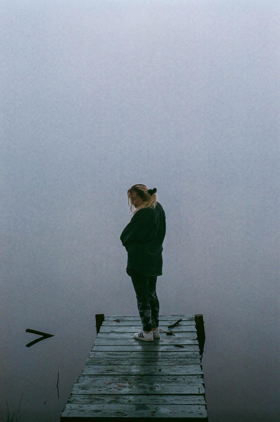 A woman stands on a wooden dock overlooking a foggy lake.