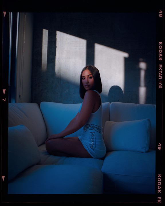 A woman sits on a white sofa with shadows from a window falling on the wall behind her.