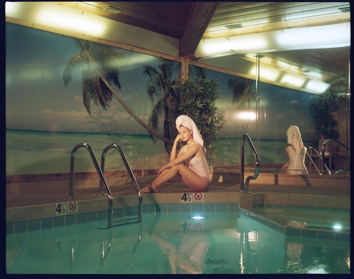 A woman in a swimsuit and towel sits by a swimming pool with a tropical mural.