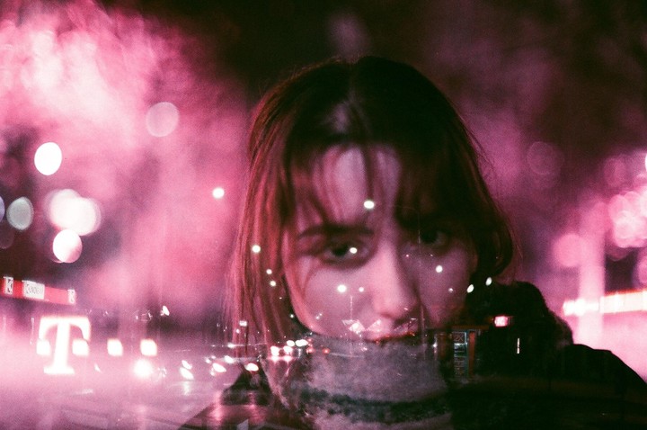 A woman is visible through a window with reflections of city lights and a pink hue.