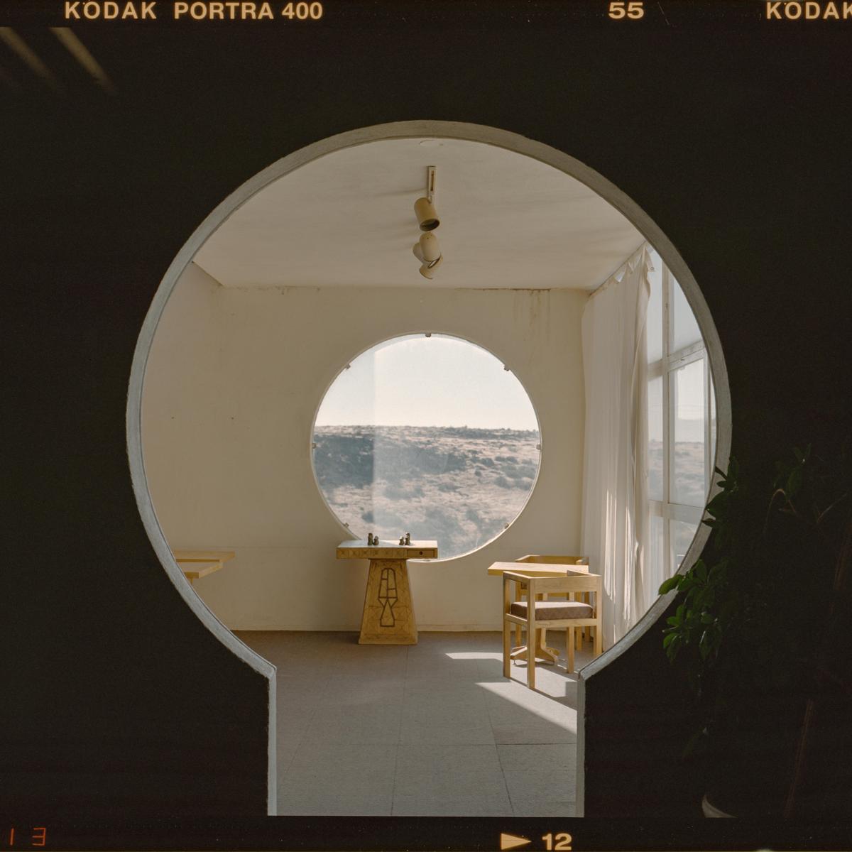 A room with a circular window overlooks a cloudy landscape, with a chess set on a pedestal and a table with chairs.