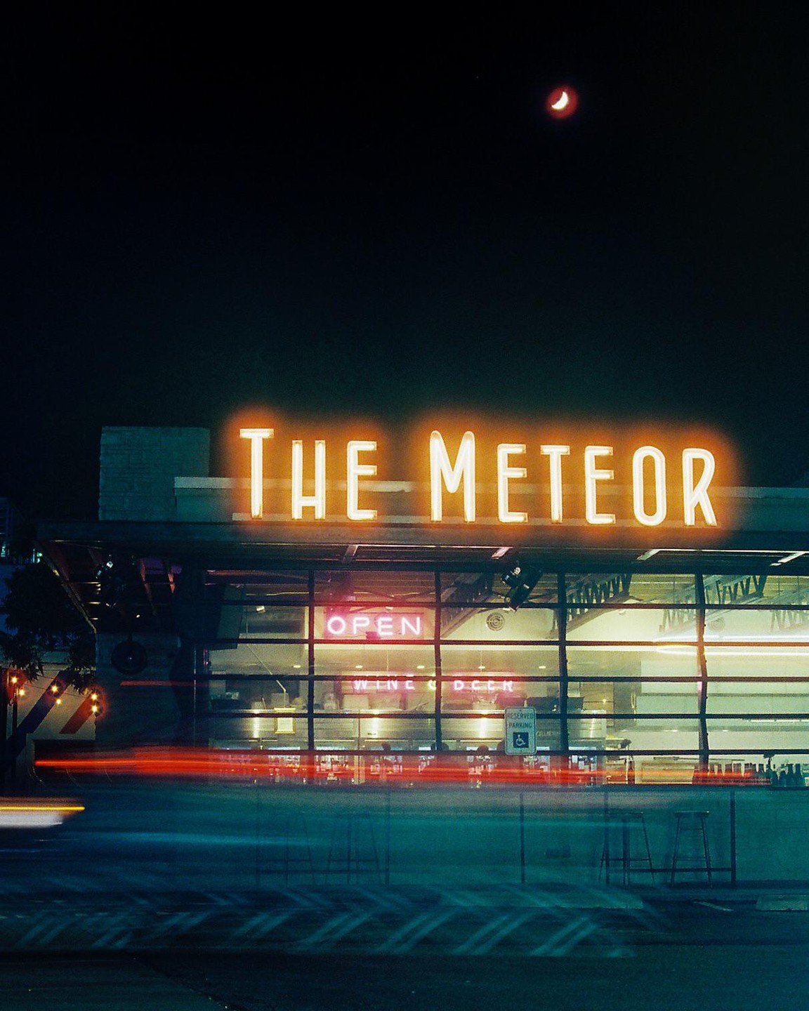 A neon sign for "THE METEOR" glows above a building with an "OPEN" sign and light trails from passing vehicles at night.