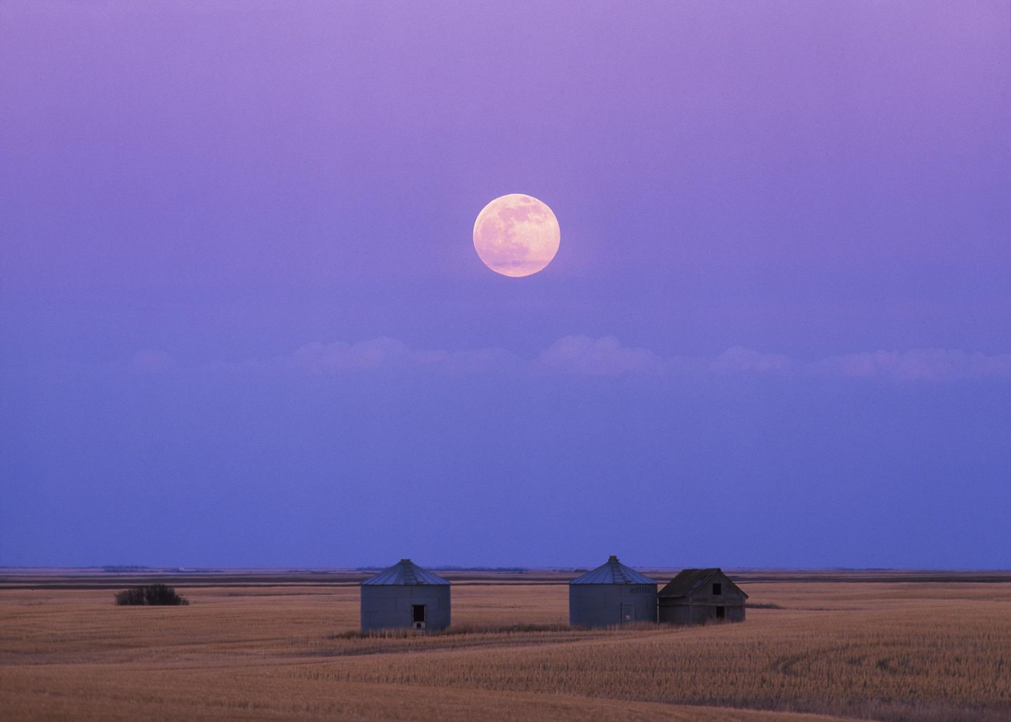 A full moon hangs in a purple sky above two grain silos and a small building in a field.