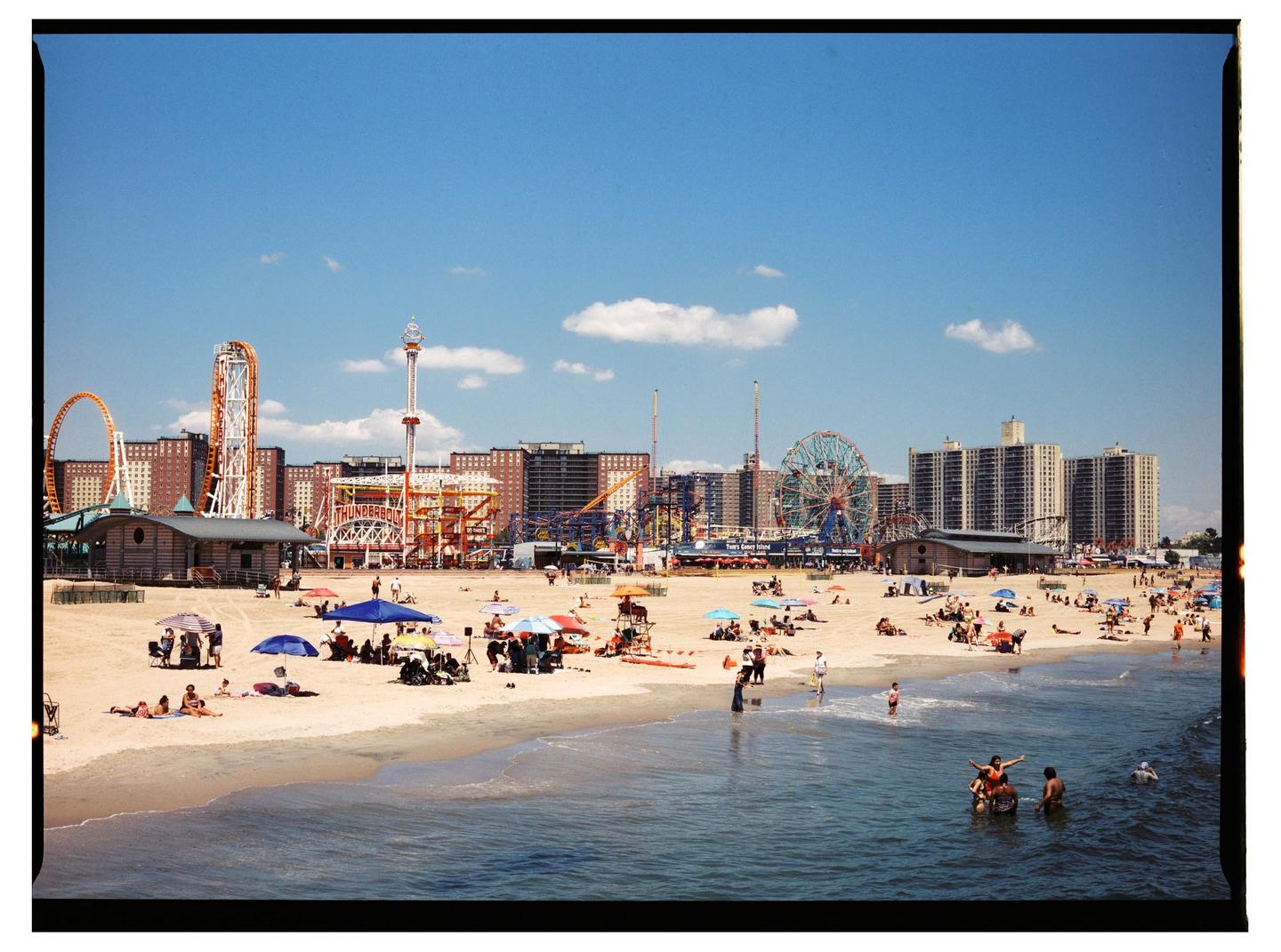 A wide shot shows a crowded beach with an amusement park and buildings in the background under a blue sky.