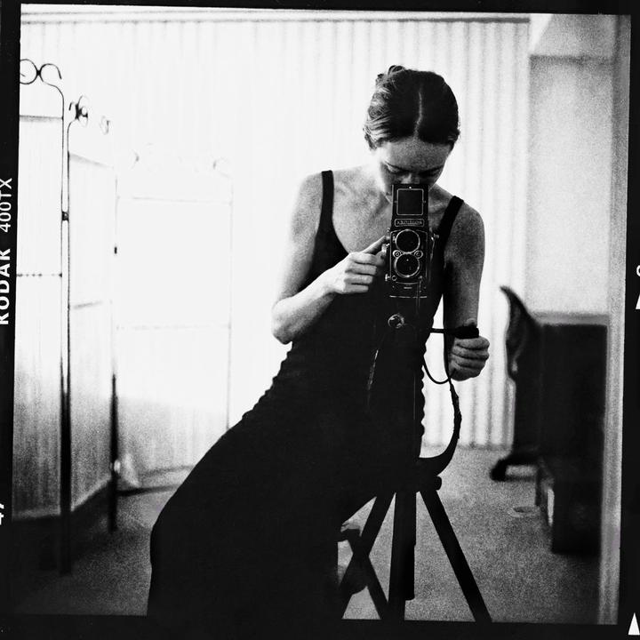 A woman in a dress photographs herself in a mirror with a camera on a tripod.