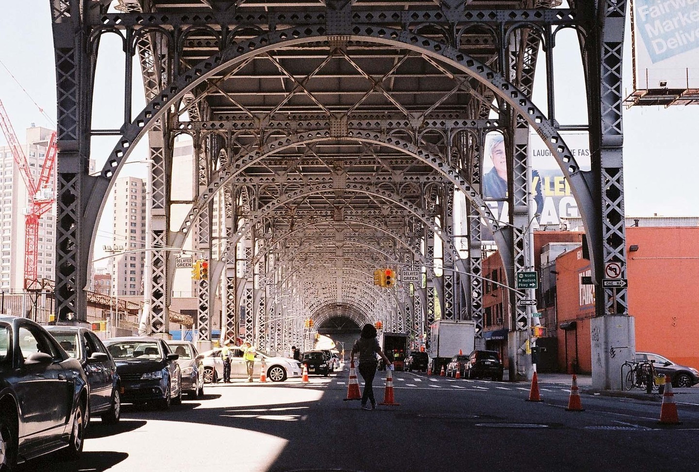 A woman walks down a street under a large metal bridge structure with cars parked on the side.