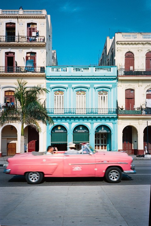 A pink vintage car with two men inside drives past colorful buildings and a palm tree.