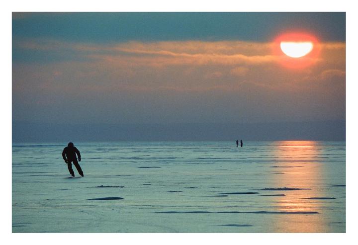 A person ice skates on a frozen lake at sunset, with two other people standing in the distance.