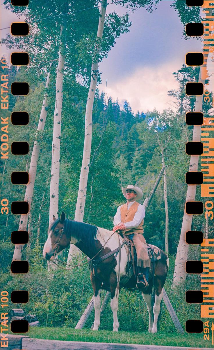 A man in a cowboy hat and vest sits on a horse in a forest of tall, slender trees.