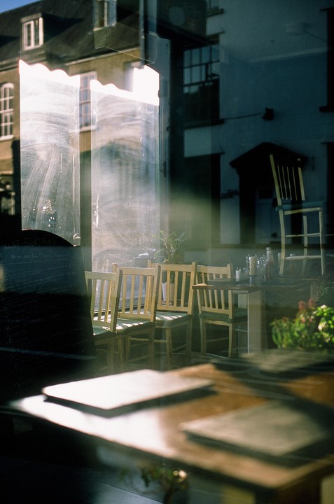 A cafe interior with wooden chairs and tables is visible through a window reflecting a building and the sky.