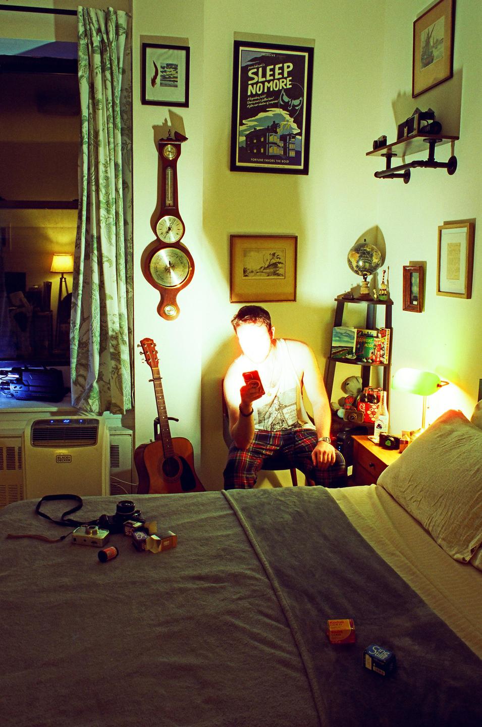A man sits in a bedroom at night, illuminated by the light of his phone.