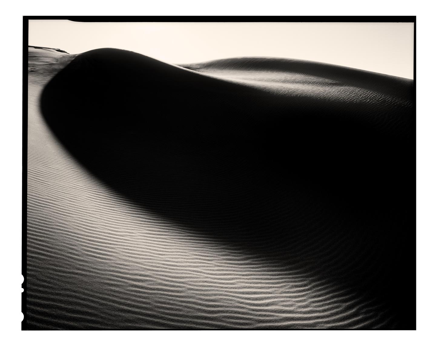 A large sand dune casts a shadow over rippled sand in a desert landscape.