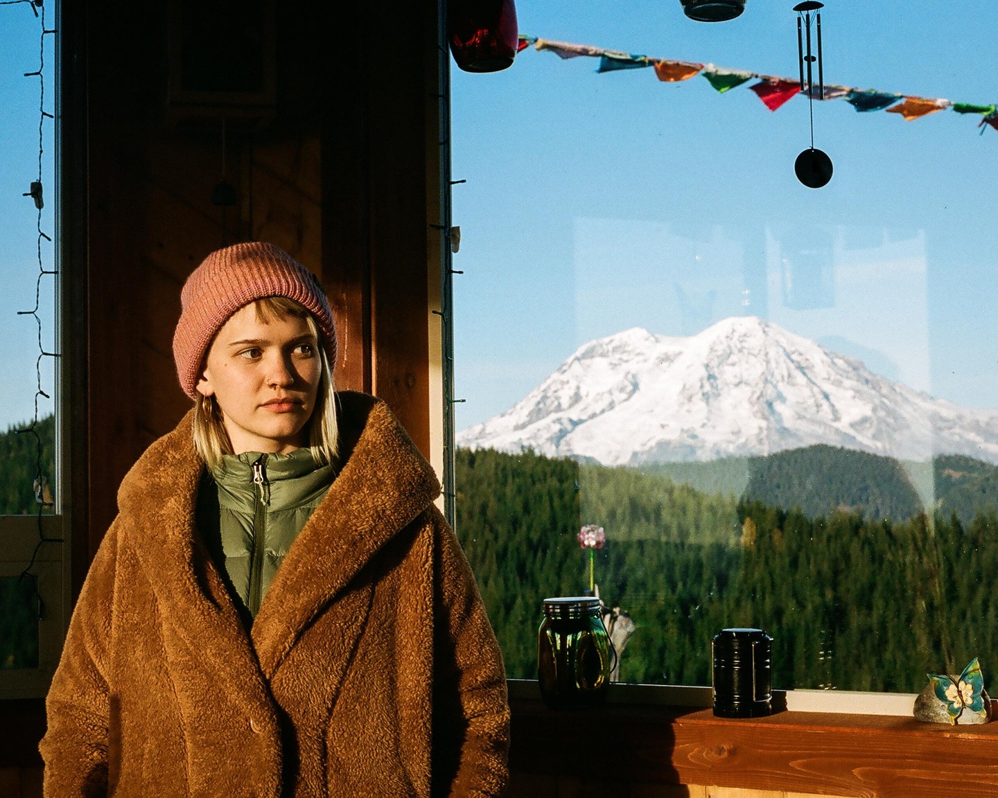 A woman in a pink beanie and brown coat stands in front of a window overlooking a snow-capped mountain and forest.