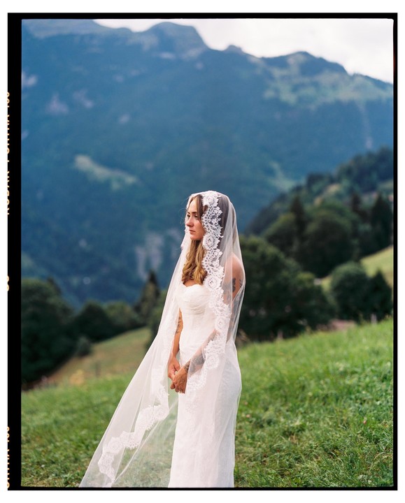 A woman in a white wedding dress and veil stands in a grassy field with mountains in the background.