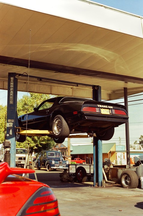 A black Trans Am is elevated on a lift at an auto repair shop, with other vehicles and tools visible around it.