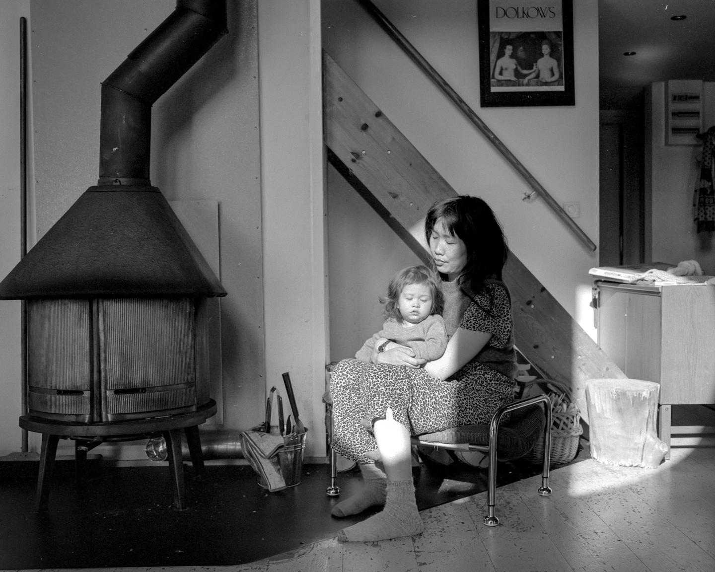 A woman holds a sleeping child on her lap in a room with a wood-burning stove and a staircase.